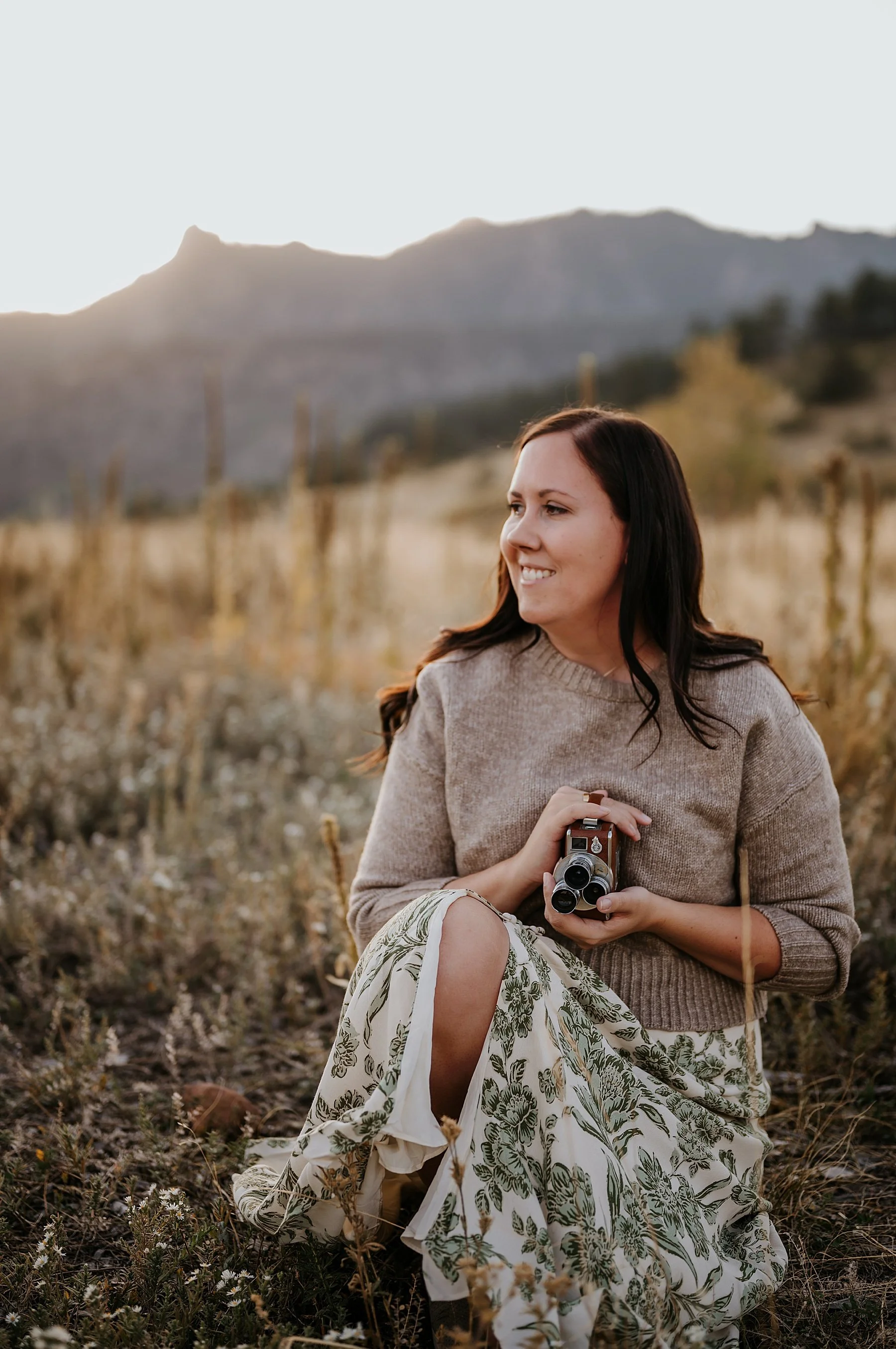 Molly sitting in a golden grass meadow holding a vintage film movie camera, with a backlit mountain ridge behind her