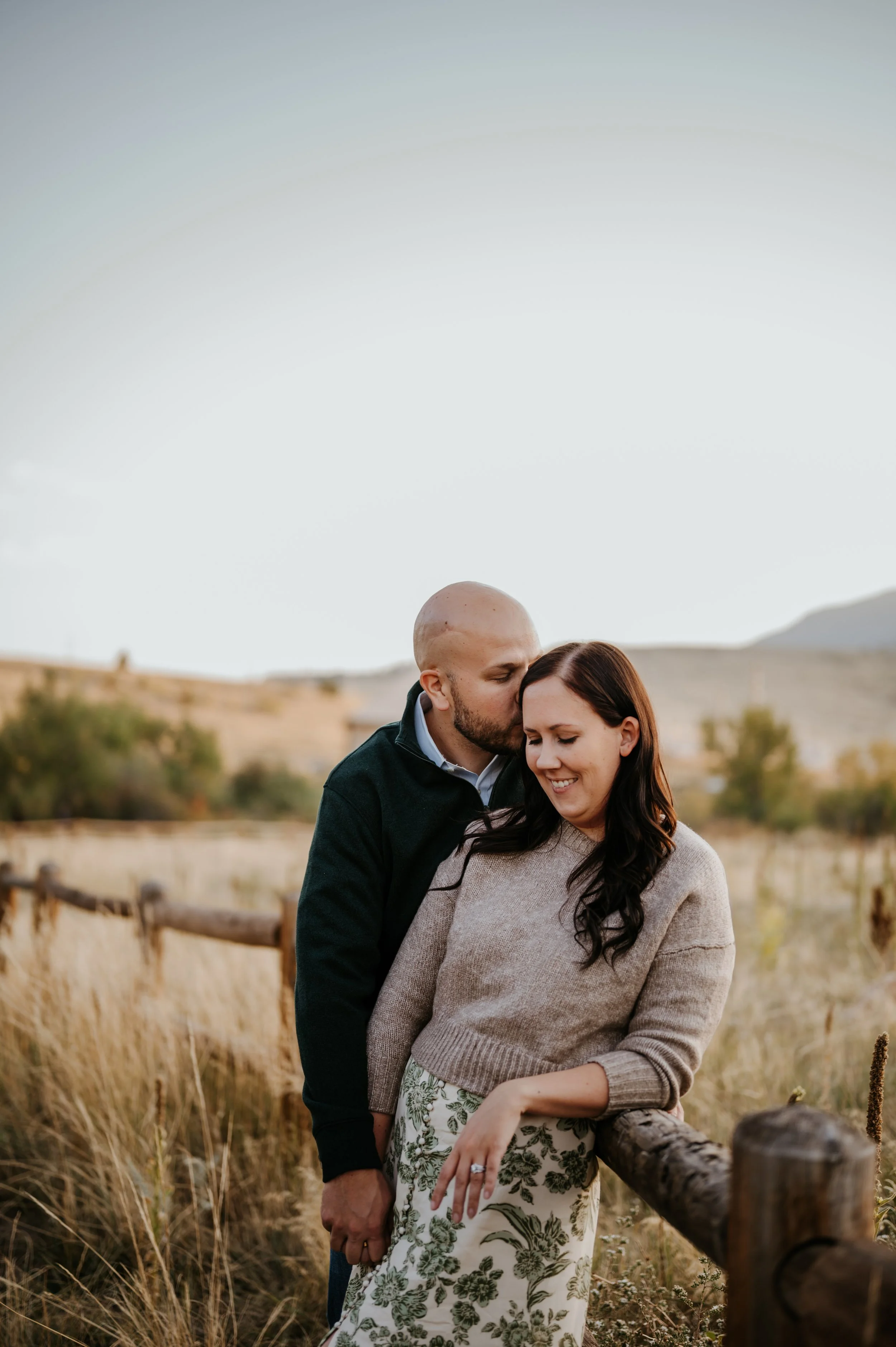 Molly leaning back against her husband beside a wooden fence in tall golden grass as he kisses the side of her head